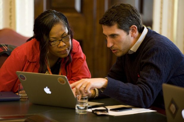 A man and a woman are looking at a laptop screen. The man is pointing at something on the screen